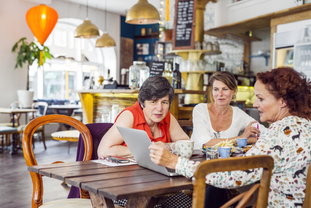 Three small business owners reviewing sponsorship licence requirements on a laptop in a café setting, discussing how to hire overseas staff.