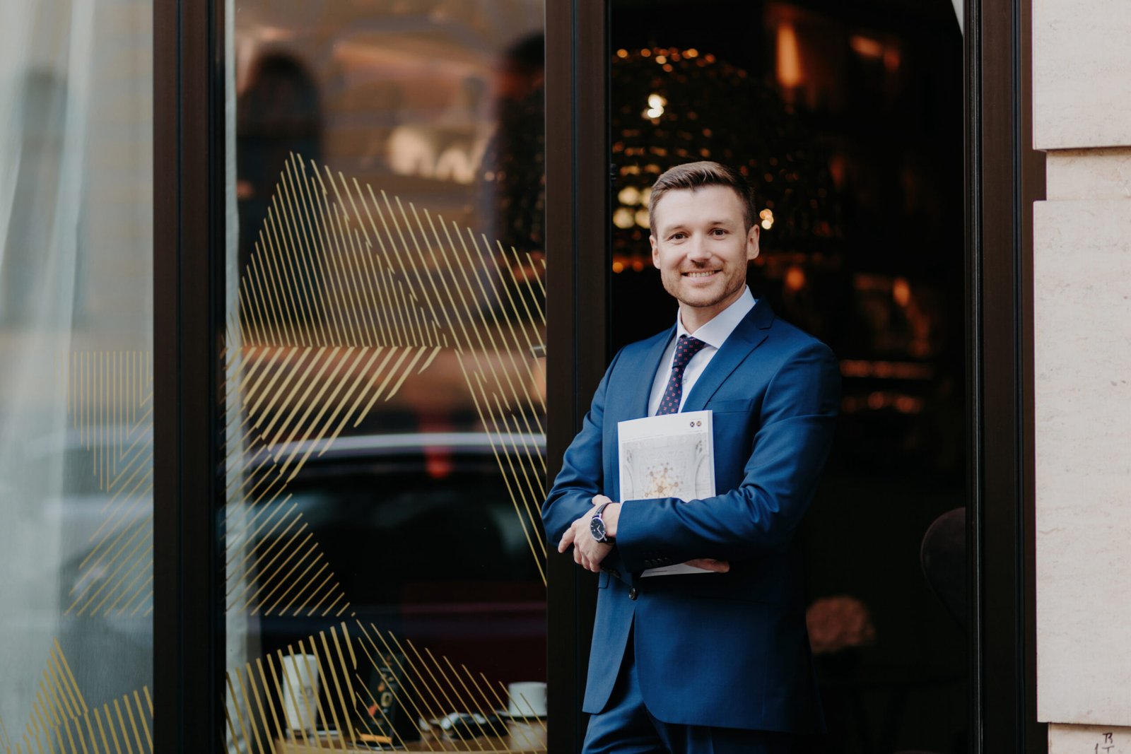 Smiling businessman in a blue suit holding documents, standing confidently outside an office building — representing guidance on whether a sponsor licence can be transferred to a new company owner.